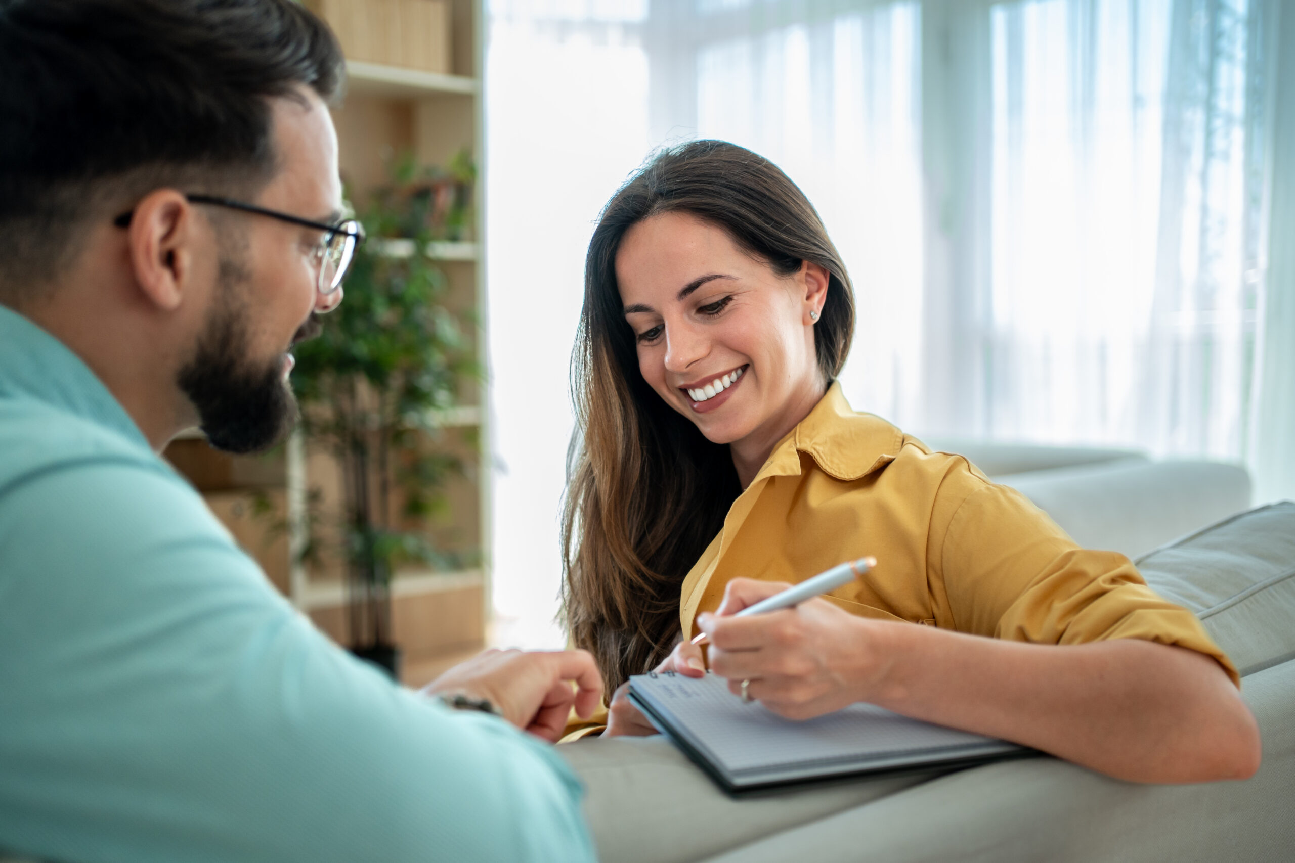 Young woman is smiling while taking notes in a notebook, sitting on a comfortable sofa next to a man, possibly during a therapy session or business meeting