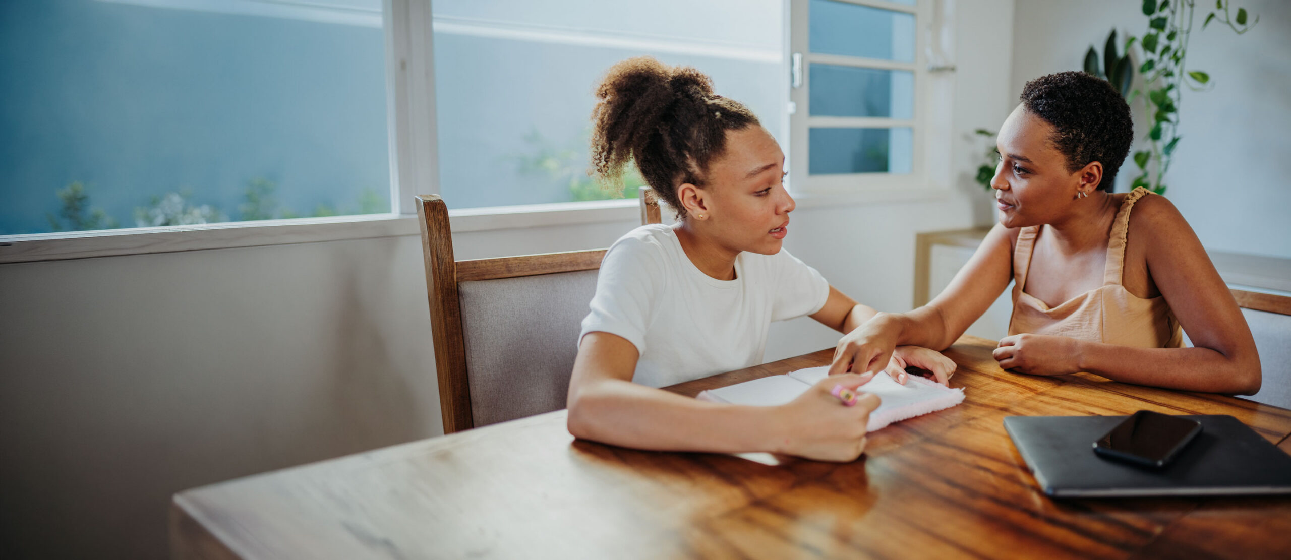 Two young women sit at a wooden table, engaged in conversation. One writes in a notebook while the other gestures supportively. A laptop and phone are on the table, and plants are visible in the background.